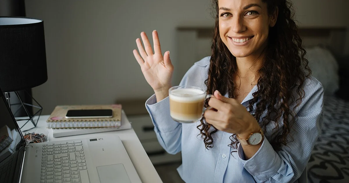 A young woman with curly hair smiles and waves while holding a cup of coffee. She sits at a desk with a laptop, notebook, and phone, creating a relaxed home office atmosphere.