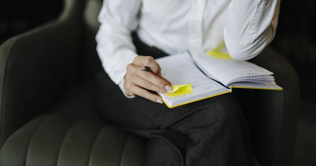 A person is sitting in a chair with a notebook and pen. They are writing notes and have sticky notes on the pages. The setting is a modern office during daylight.
